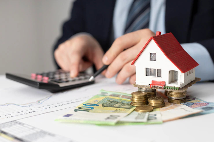 Fundo Imobiliário: uma pessoa faz contas na calculadora que está numa mesa branca, próximo a maquete de uma casa pequena e várias moedas e notas de dinheiro.
